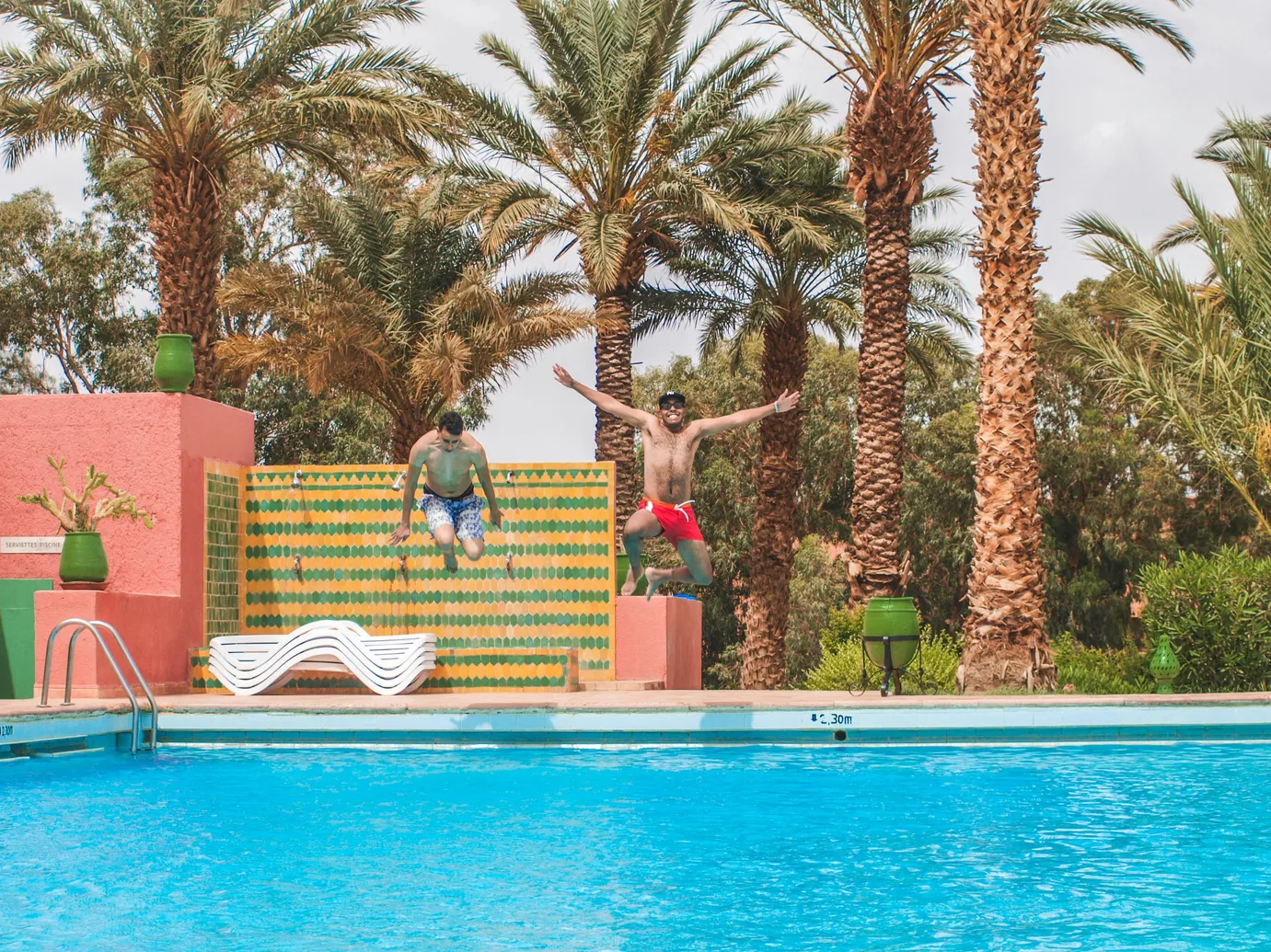 Two people jumping into a fiberglass pool surrounded by palm trees, illustrating the joy of owning a fiberglass pool.